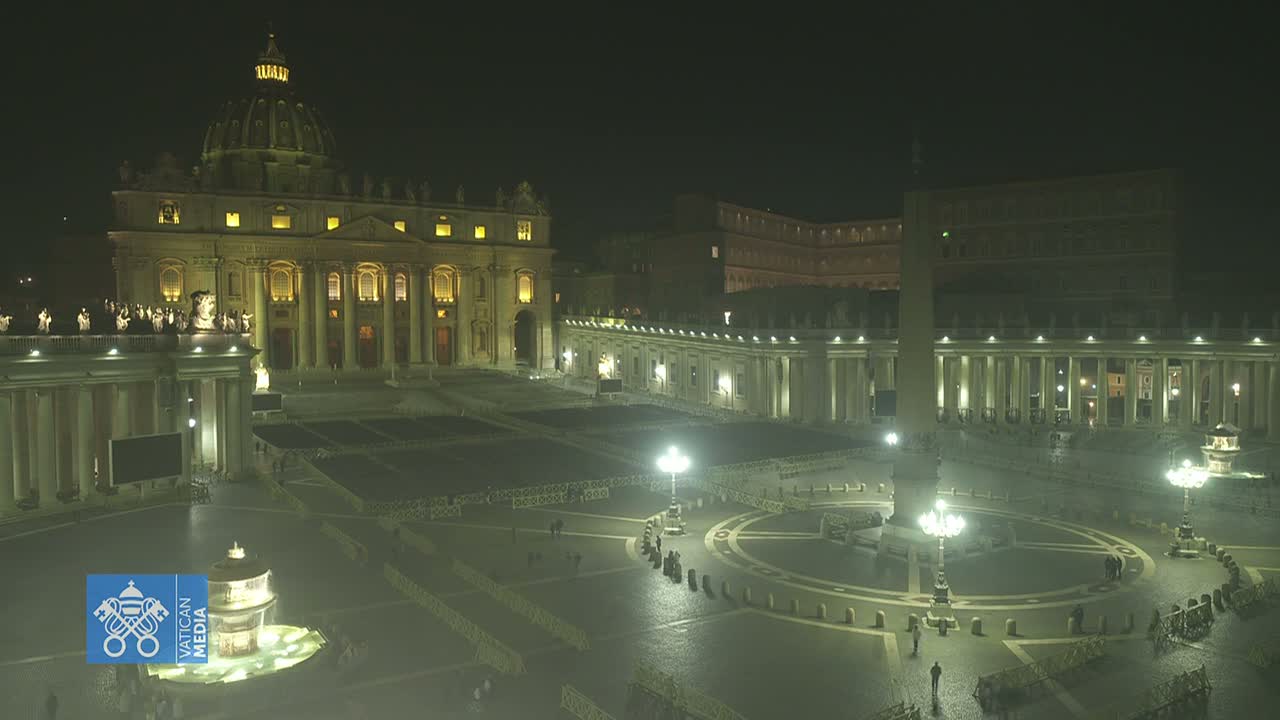 St. Peter's Basilica and Square are illuminated at night, with a few people visible in the vast, mostly empty space.