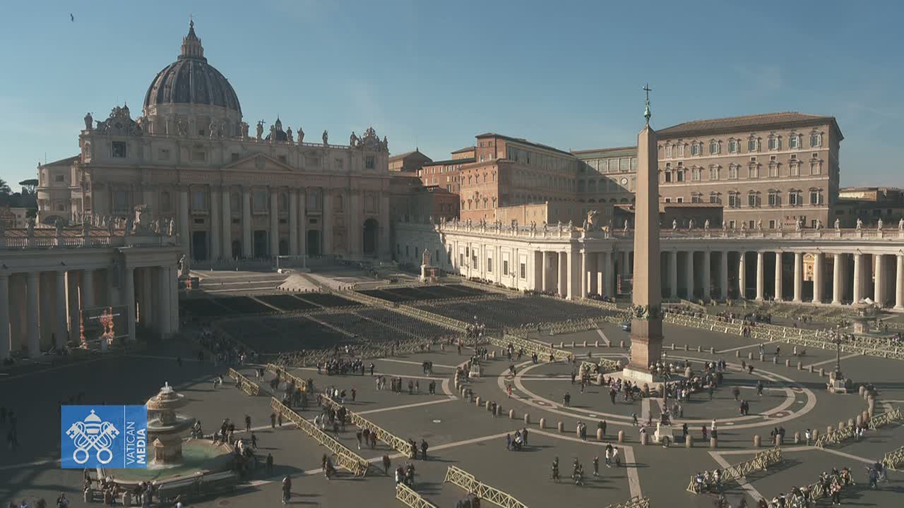 St. Peter's Square in Vatican City, under a clear sunny sky, shows many people gathered around a large obelisk in the center of the plaza, with St. Peter's Basilica in the background.