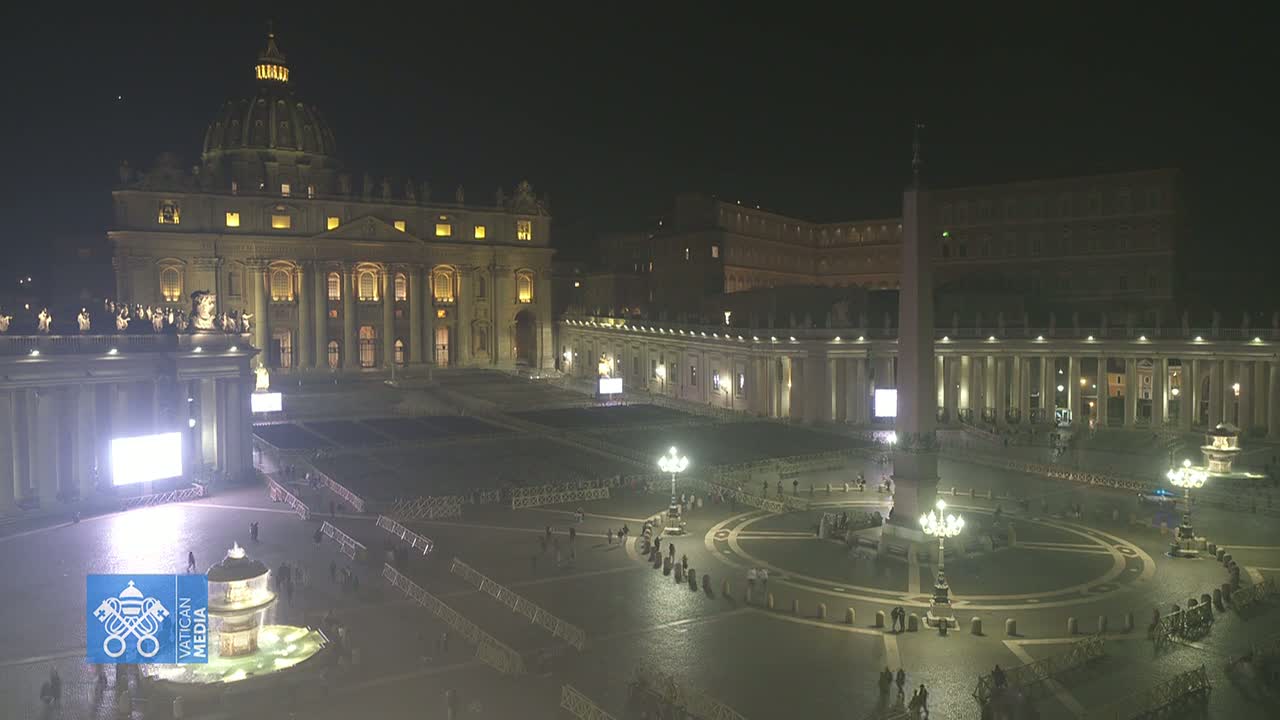 At night, St. Peter's Basilica and Square are sparsely populated, illuminated, and show a large screen and barriers in the square.