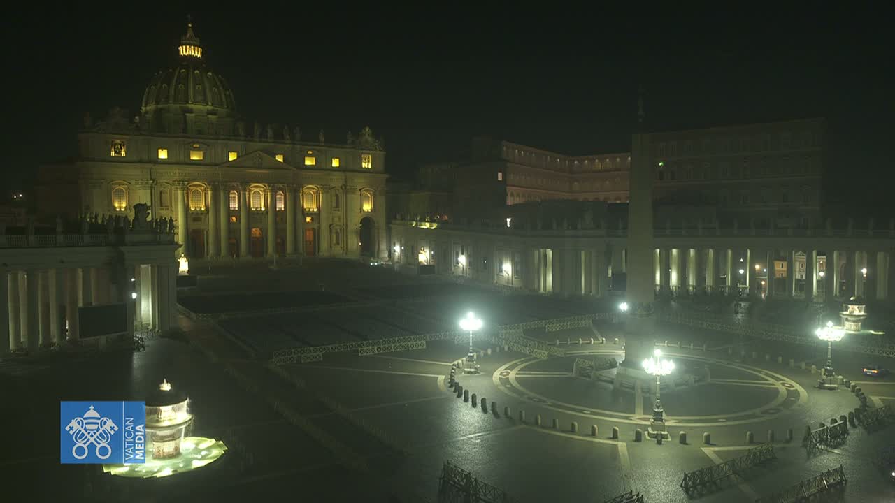 A nighttime, dimly lit view of St. Peter's Basilica and Square, showing a mostly empty plaza with some lights and a fountain.