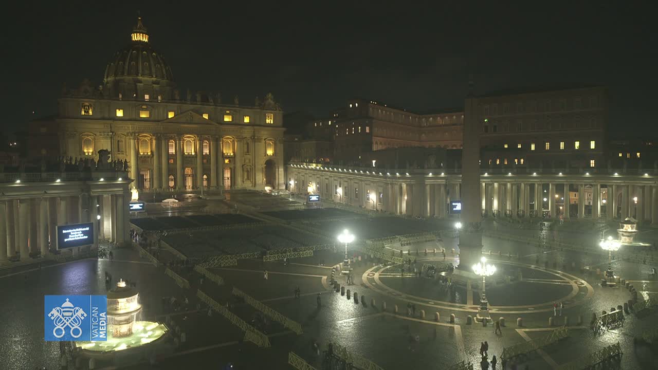 St. Peter's Basilica and St. Peter's Square at night, illuminated, with a wet, empty plaza and a few people scattered throughout.