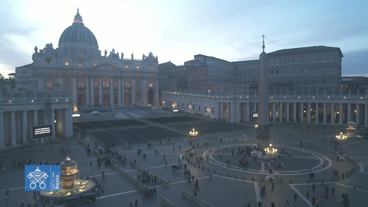 St. Peter's Square in Vatican City at dusk, showing St. Peter's Basilica and the surrounding colonnades, with a few people scattered throughout the large plaza.