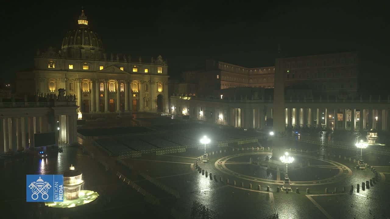 A nighttime, wet view of St. Peter's Basilica and St. Peter's Square, illuminated, with many rows of chairs set up in the square.