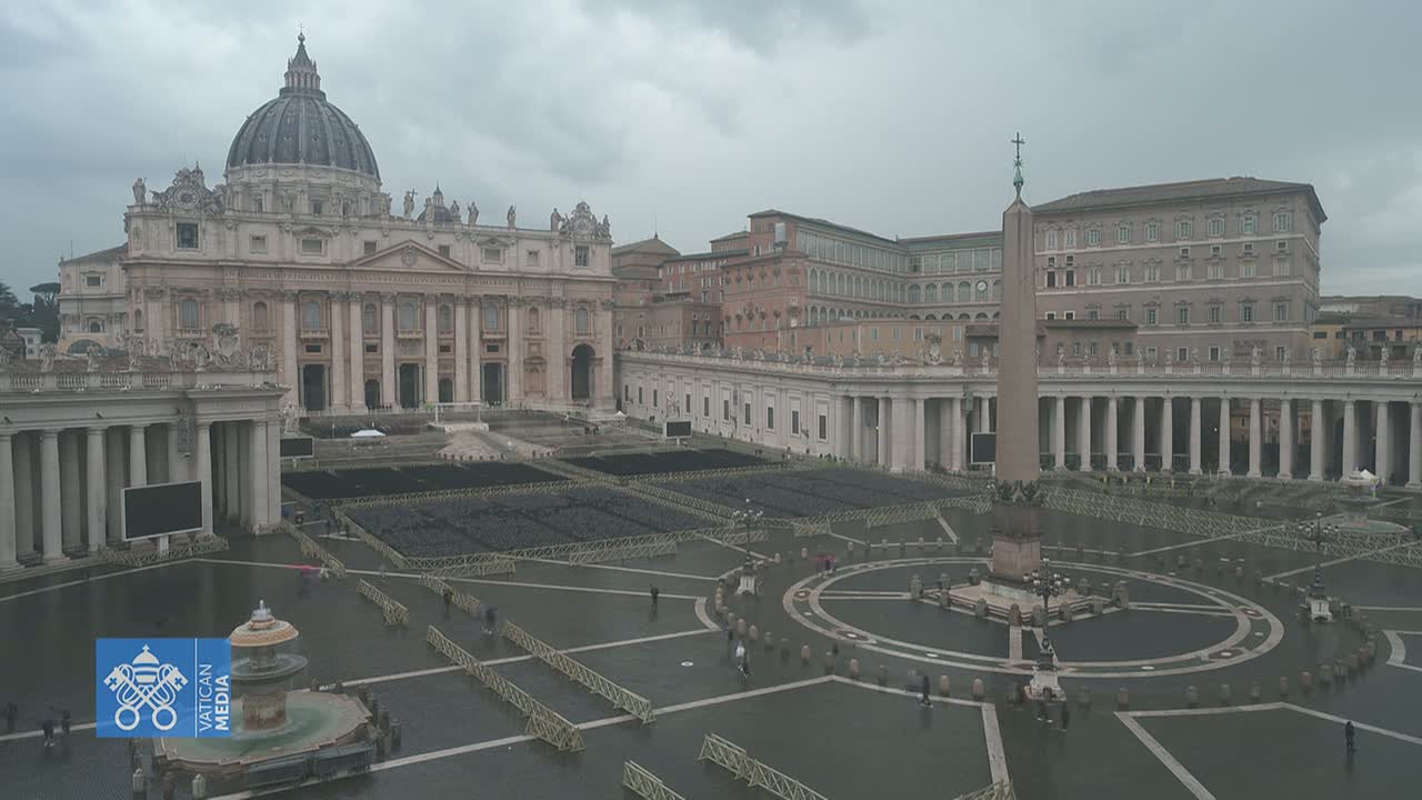 St. Peter's Basilica and Square in Vatican City are shown on an overcast day, with the square largely empty except for some people and what appears to be preparations for an event.
