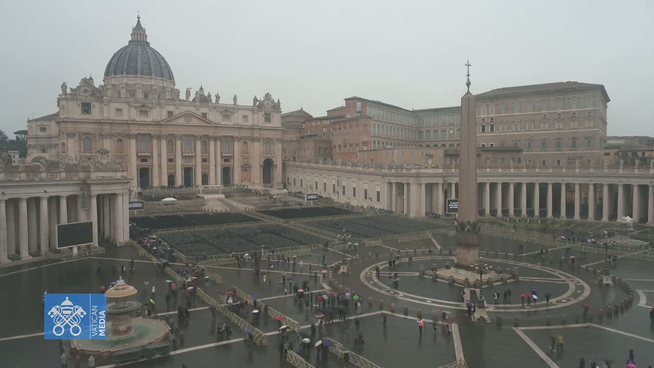 St. Peter's Basilica and St. Peter's Square in Vatican City are shown on a rainy day, with many people carrying umbrellas and walking across the wet plaza.