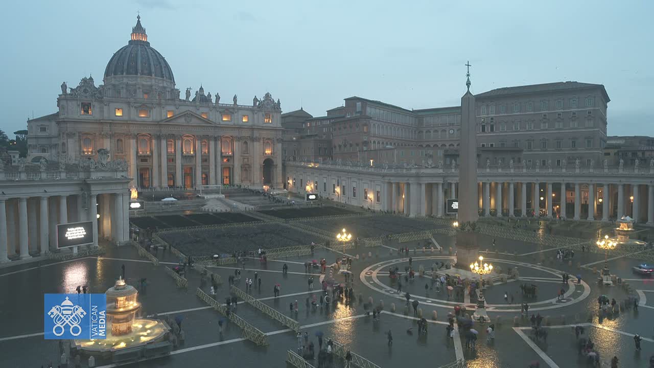 A large, illuminated basilica and plaza are shown under a gloomy, wet sky, with many people scattered throughout the wet stone square.