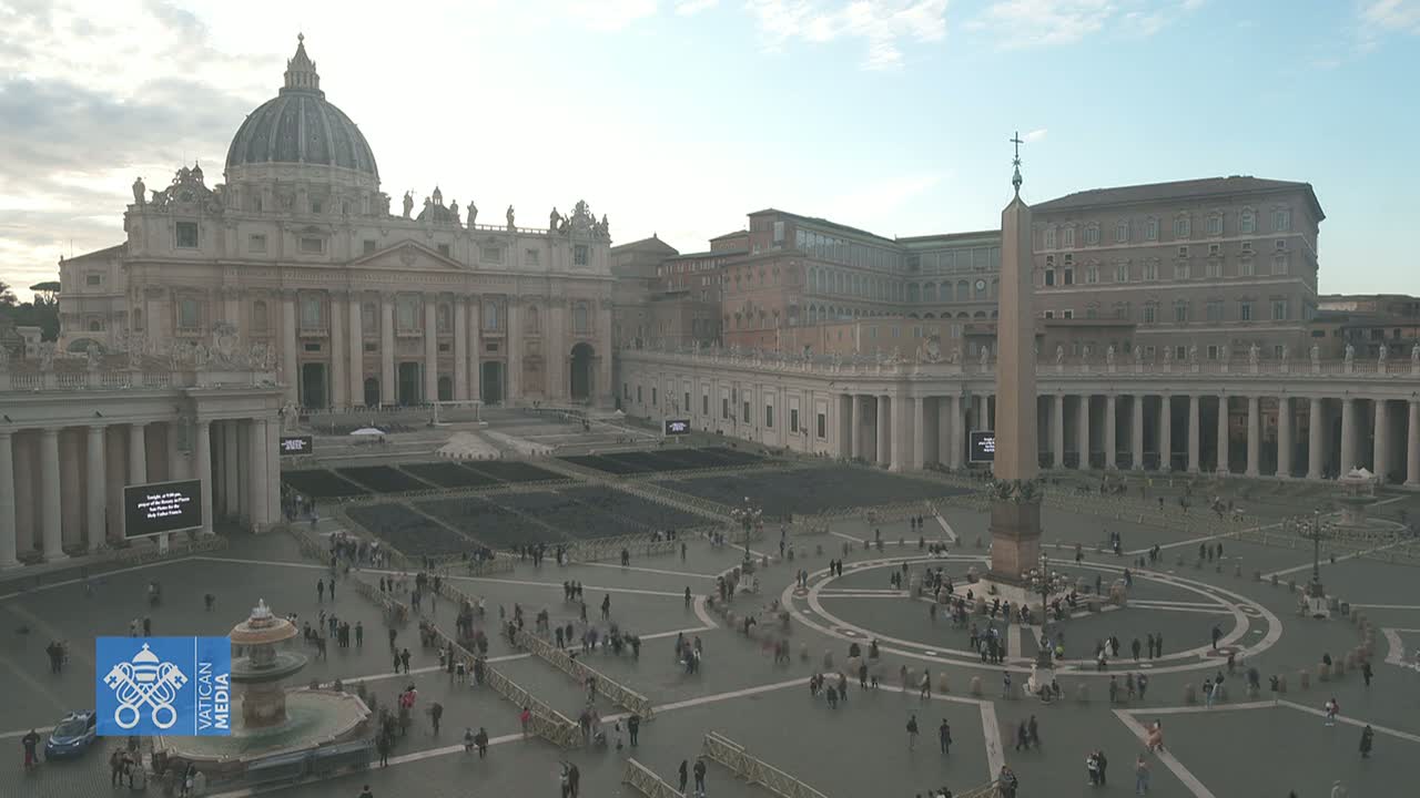 St. Peter's Square in Vatican City, under a mostly cloudy sky, shows a large crowd of people gathered around an obelisk in the center of the plaza, with St. Peter's Basilica in the background.