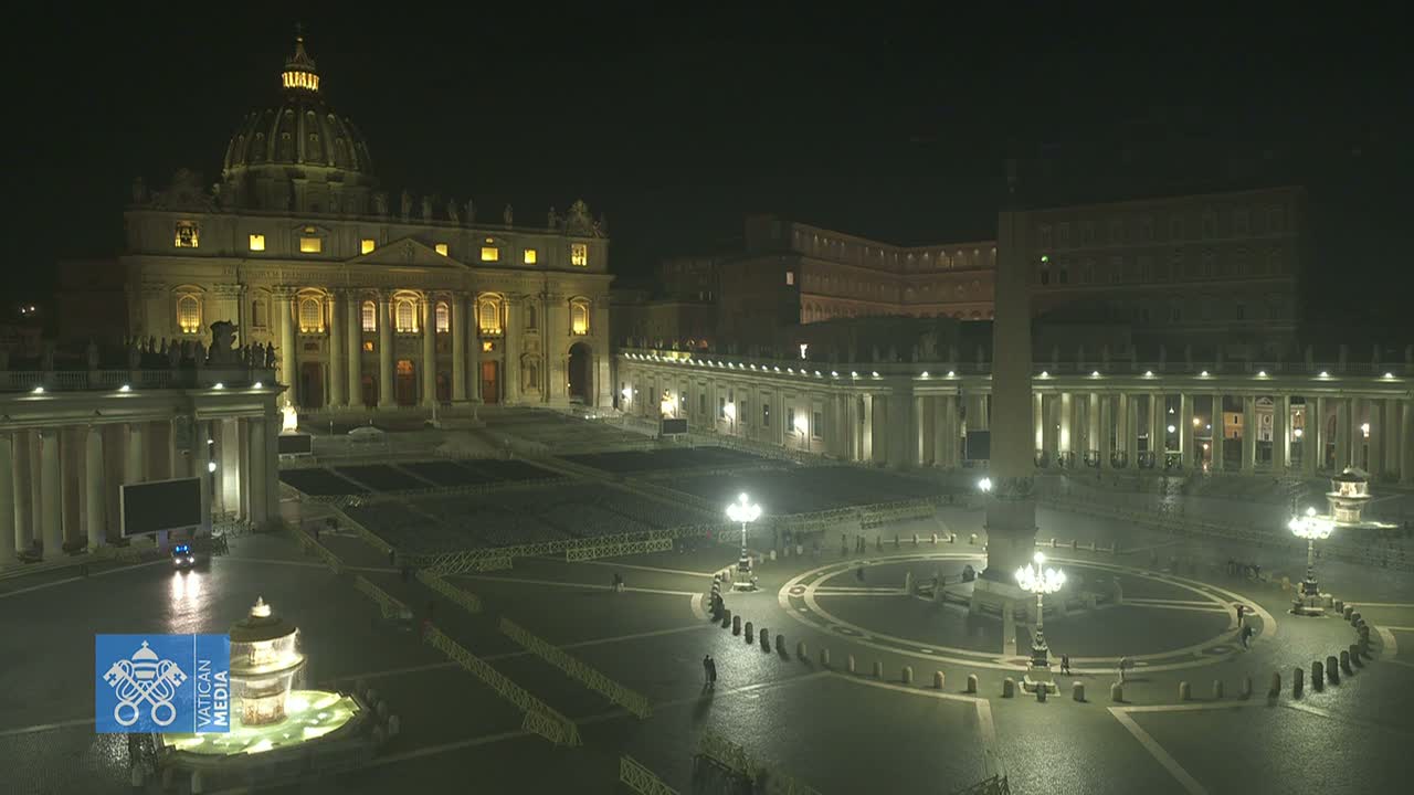 St. Peter's Basilica and Square are illuminated at night, with a mostly empty plaza except for a few people and some barriers.
