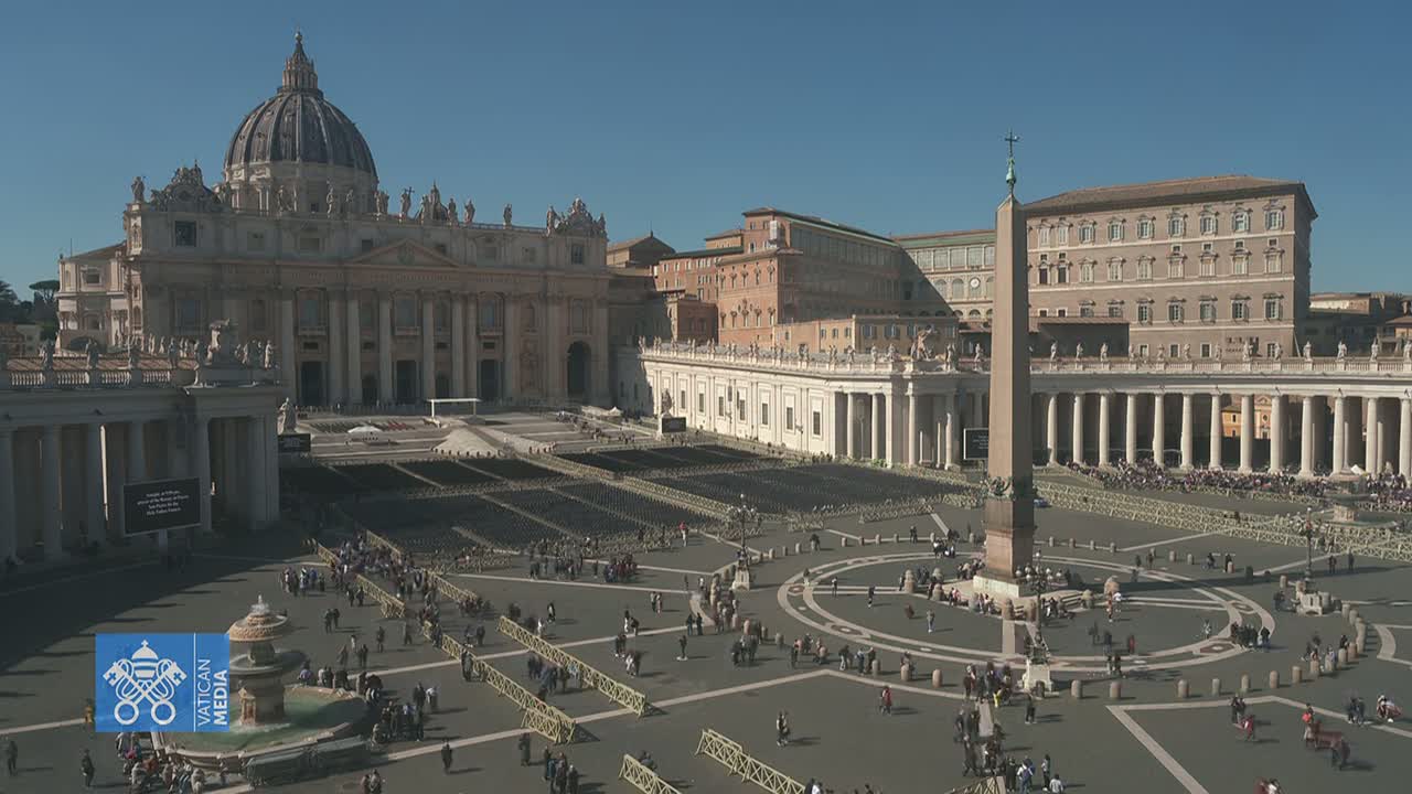 St. Peter's Square in Vatican City, under a clear sunny sky, is filled with people gathered in the plaza surrounding St. Peter's Basilica and an obelisk.