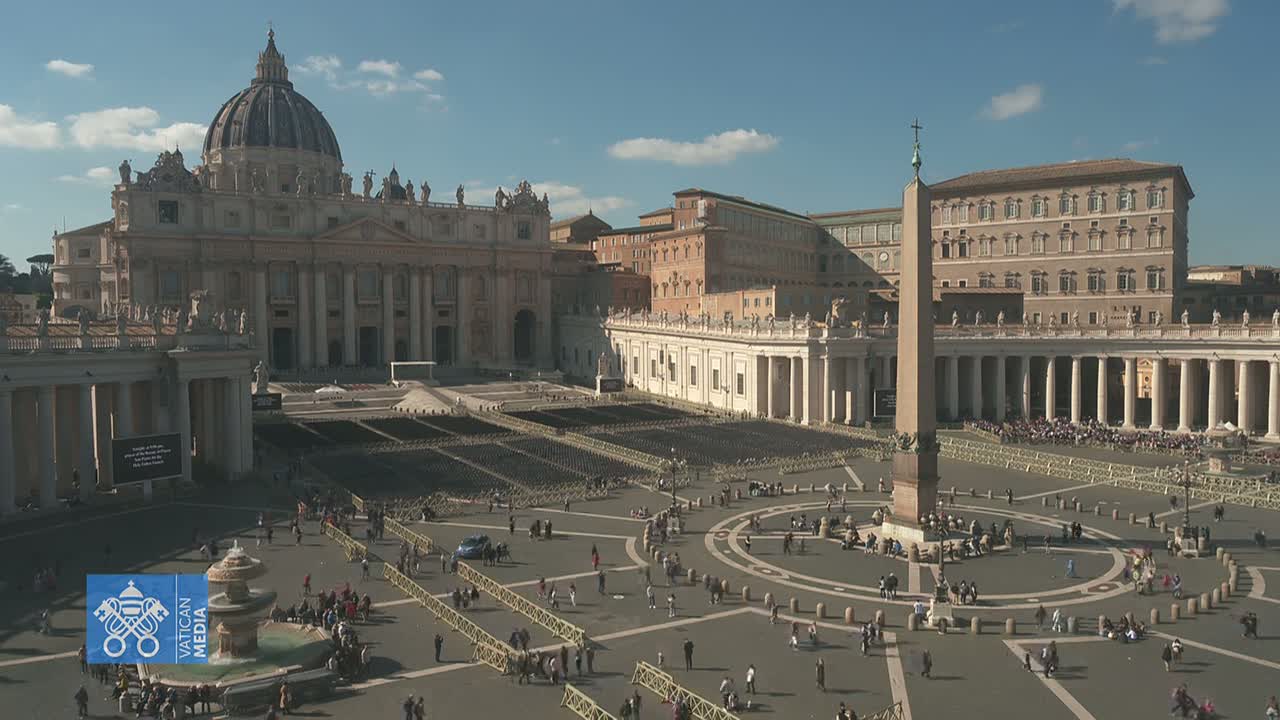 St. Peter's Square in Vatican City, under a mostly sunny sky, is filled with many people gathered around a large obelisk in front of St. Peter's Basilica.