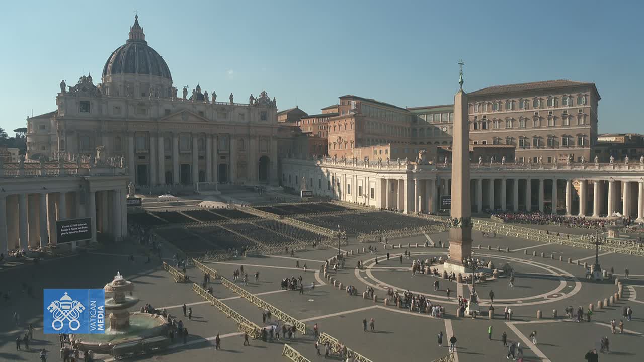 St. Peter's Square in Vatican City, under a clear, sunny sky, is mostly empty except for a few people and many rows of empty chairs set up in the square.
