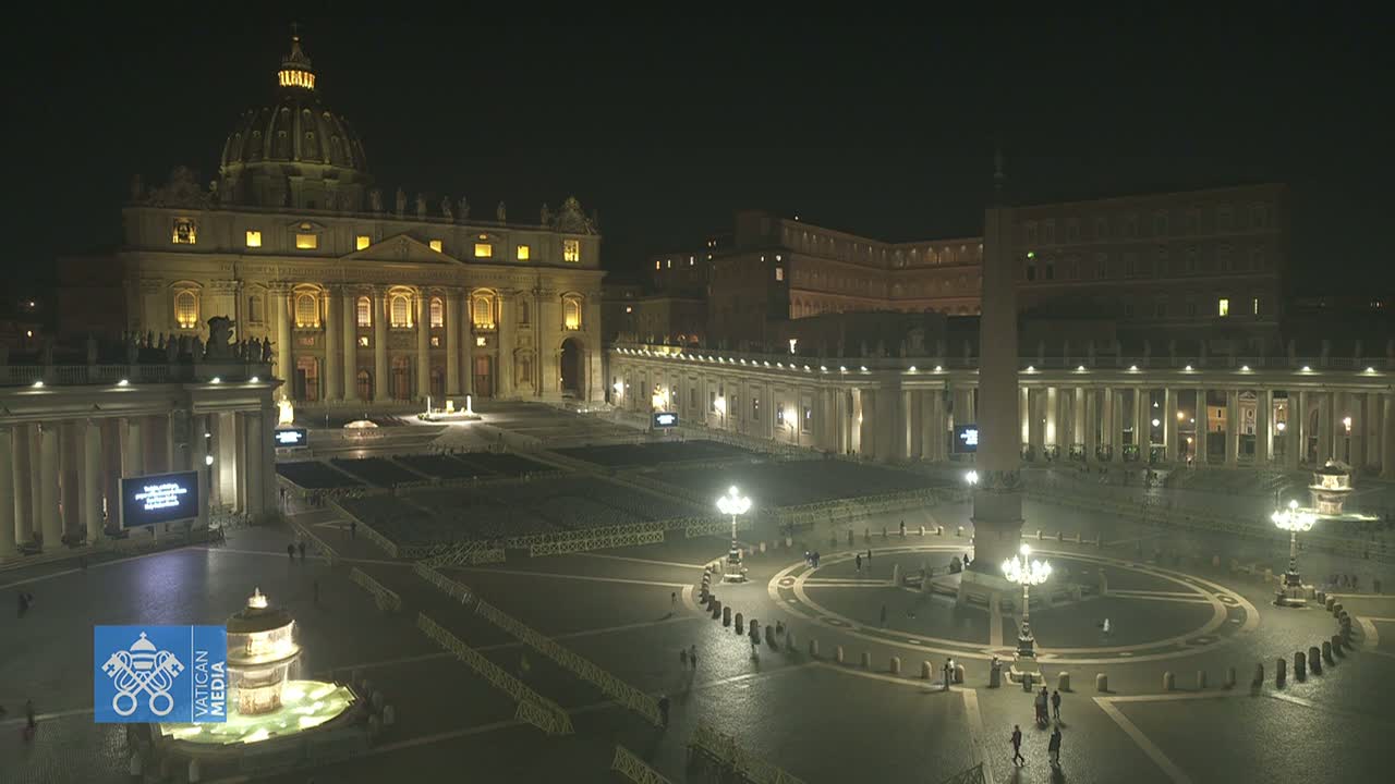 St. Peter's Basilica and Square are illuminated at night, with a large expanse of seating set up in the square, and a few people are visible walking around.