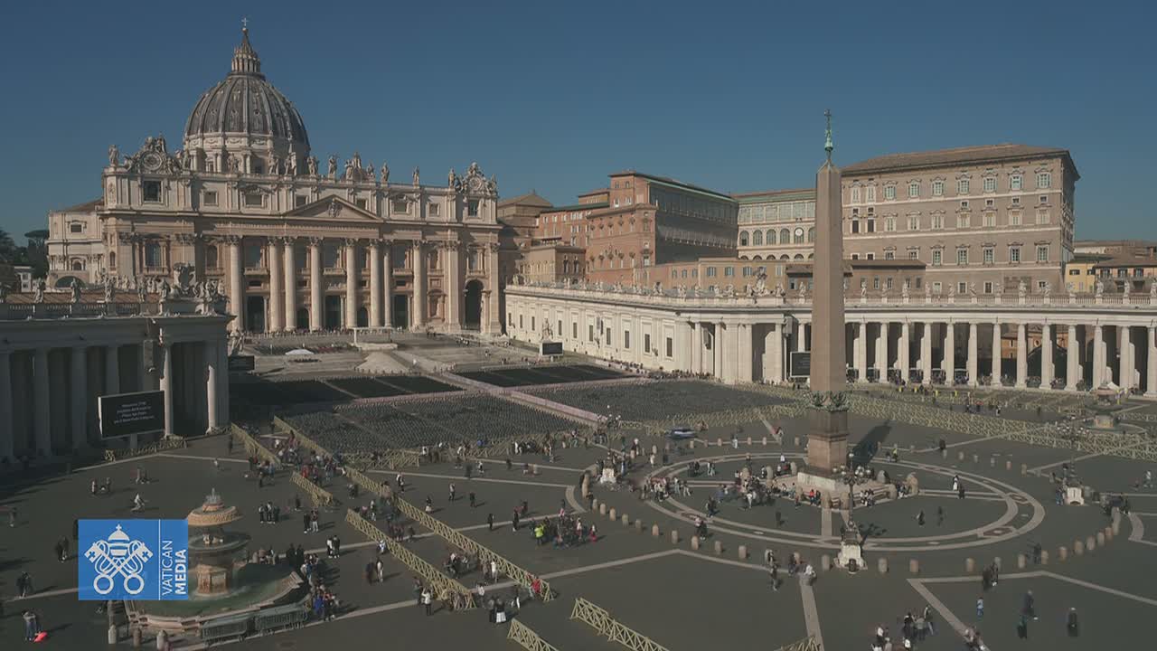 St. Peter's Basilica and St. Peter's Square in Vatican City, under a clear blue sky, are shown from an elevated perspective, with numerous people scattered throughout the square.