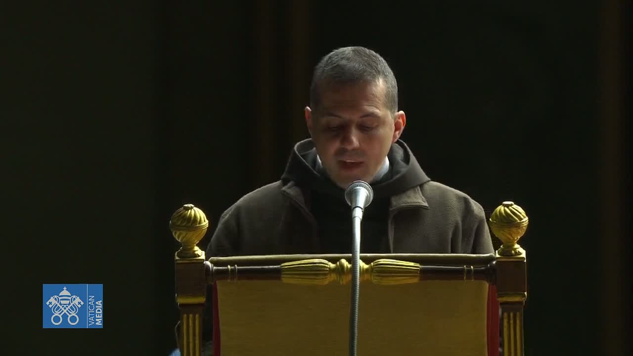 A man in a brown robe speaks into a microphone at a dark wooden lectern.