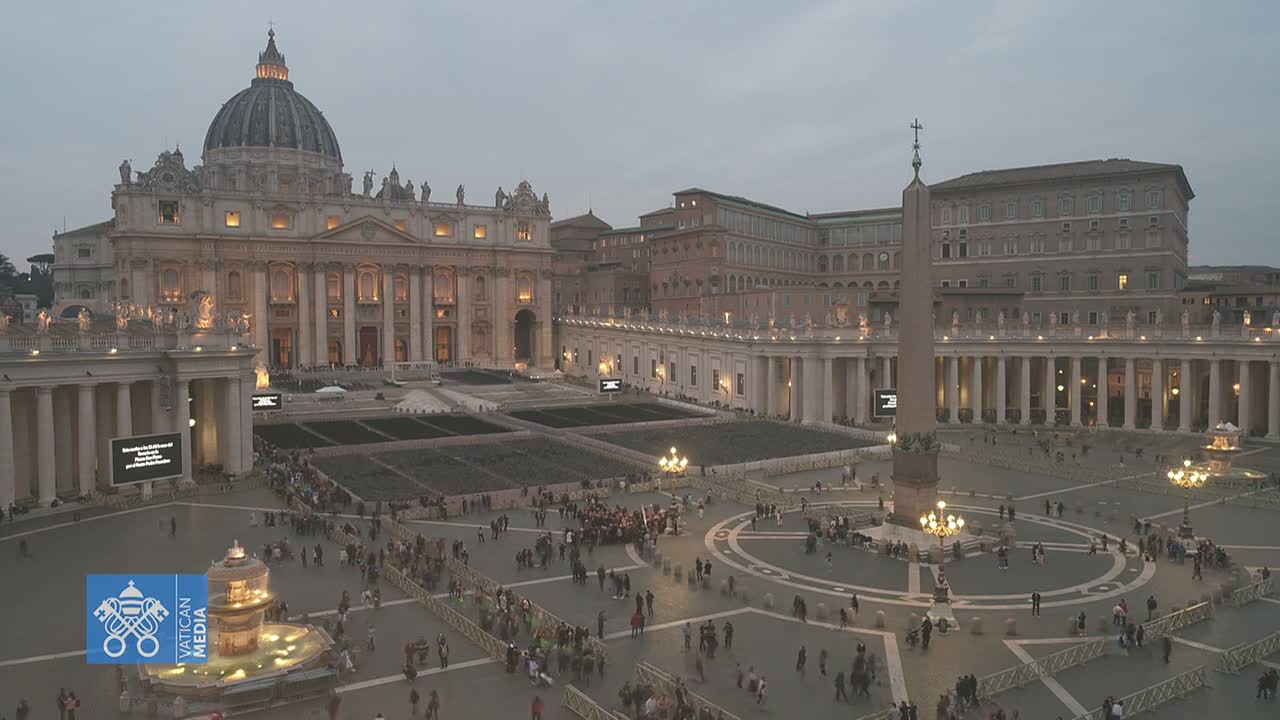 St. Peter's Basilica and St. Peter's Square are illuminated at dusk, with a crowd of people gathered in the square.