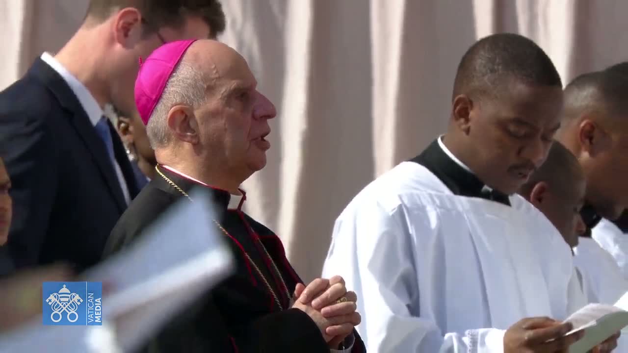 A bishop in a pink zucchetto stands with his hands clasped, surrounded by other clergy in white robes, outdoors in bright sunlight.