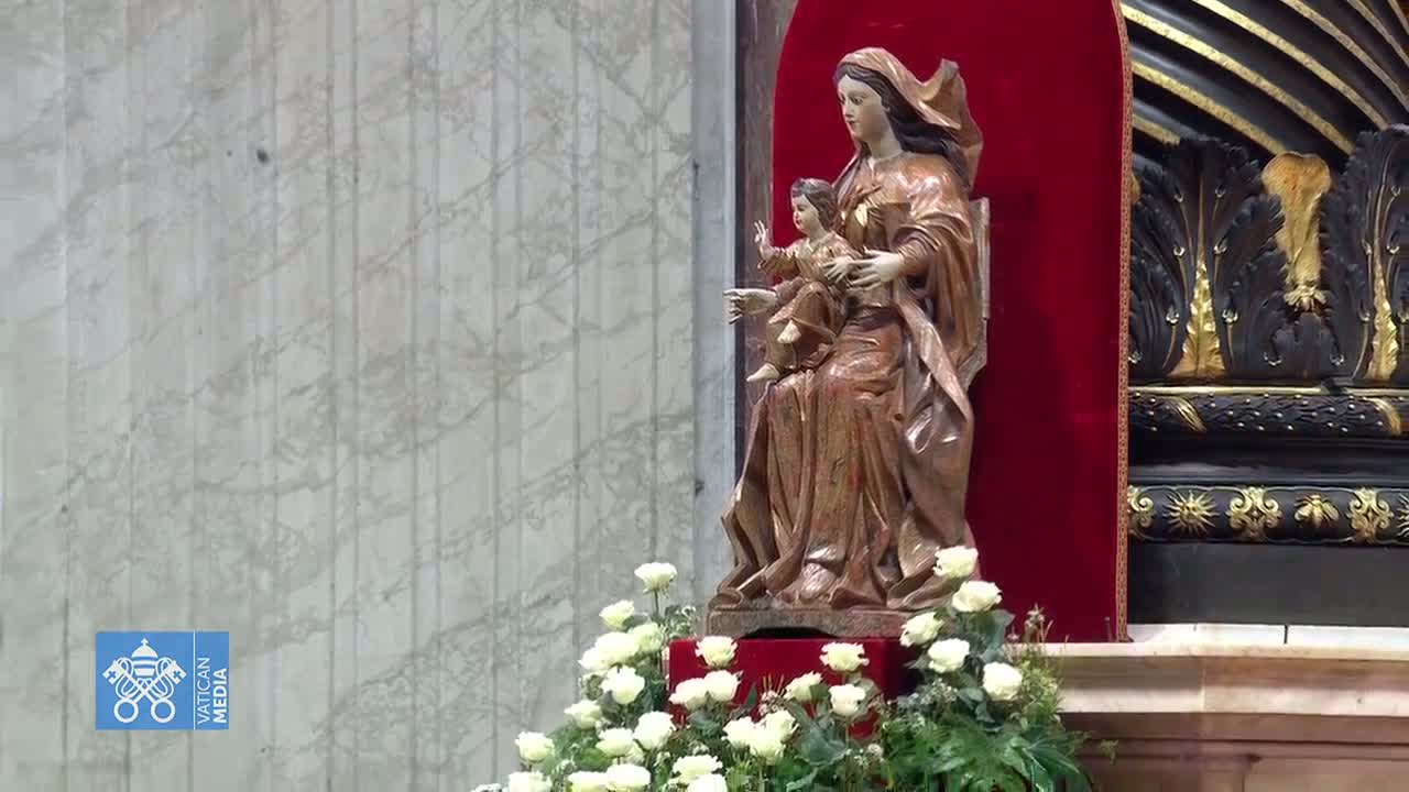 A wooden statue of Mary and the baby Jesus sits in a red alcove, adorned with white roses.