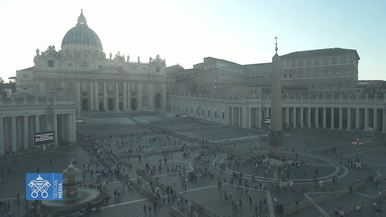 A hazy, sunlit view of St. Peter's Square in Vatican City shows many people gathered in the plaza surrounding St. Peter's Basilica and an obelisk.