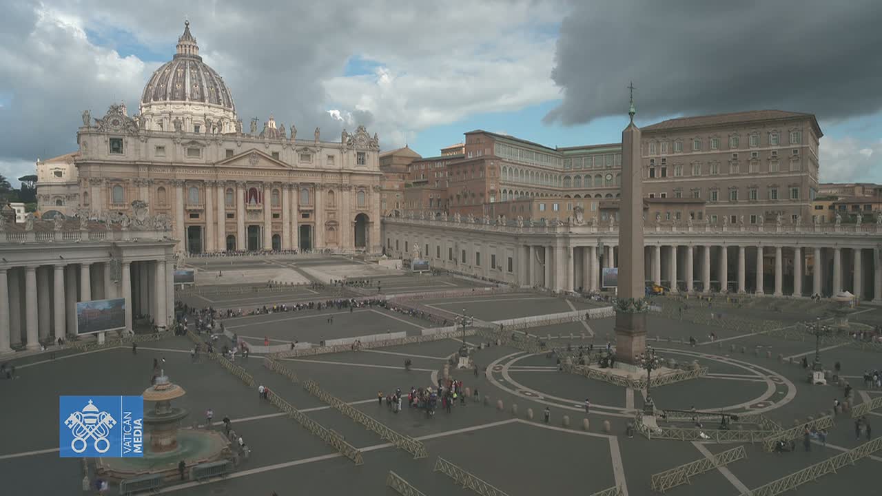 St. Peter's Square in Vatican City, under a cloudy sky, shows the basilica and surrounding buildings with a few people scattered throughout the large plaza.