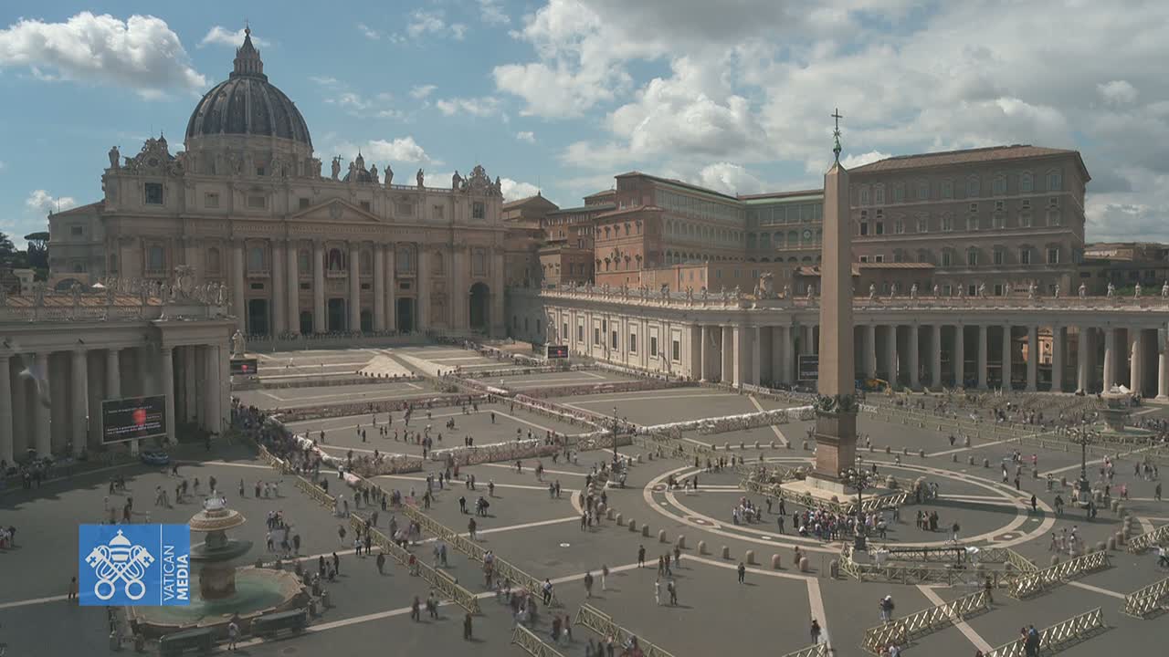 St. Peter's Basilica and Square are shown on a partly cloudy day, with many people gathered in the square, which is sectioned off with barriers.