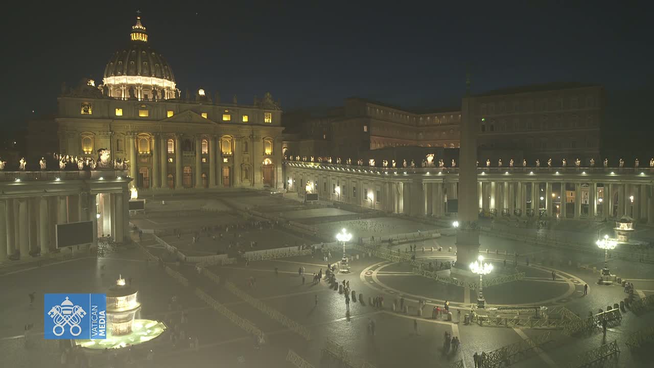 A brightly lit St. Peter's Basilica and Square at night, with a few people sparsely scattered throughout the large plaza.