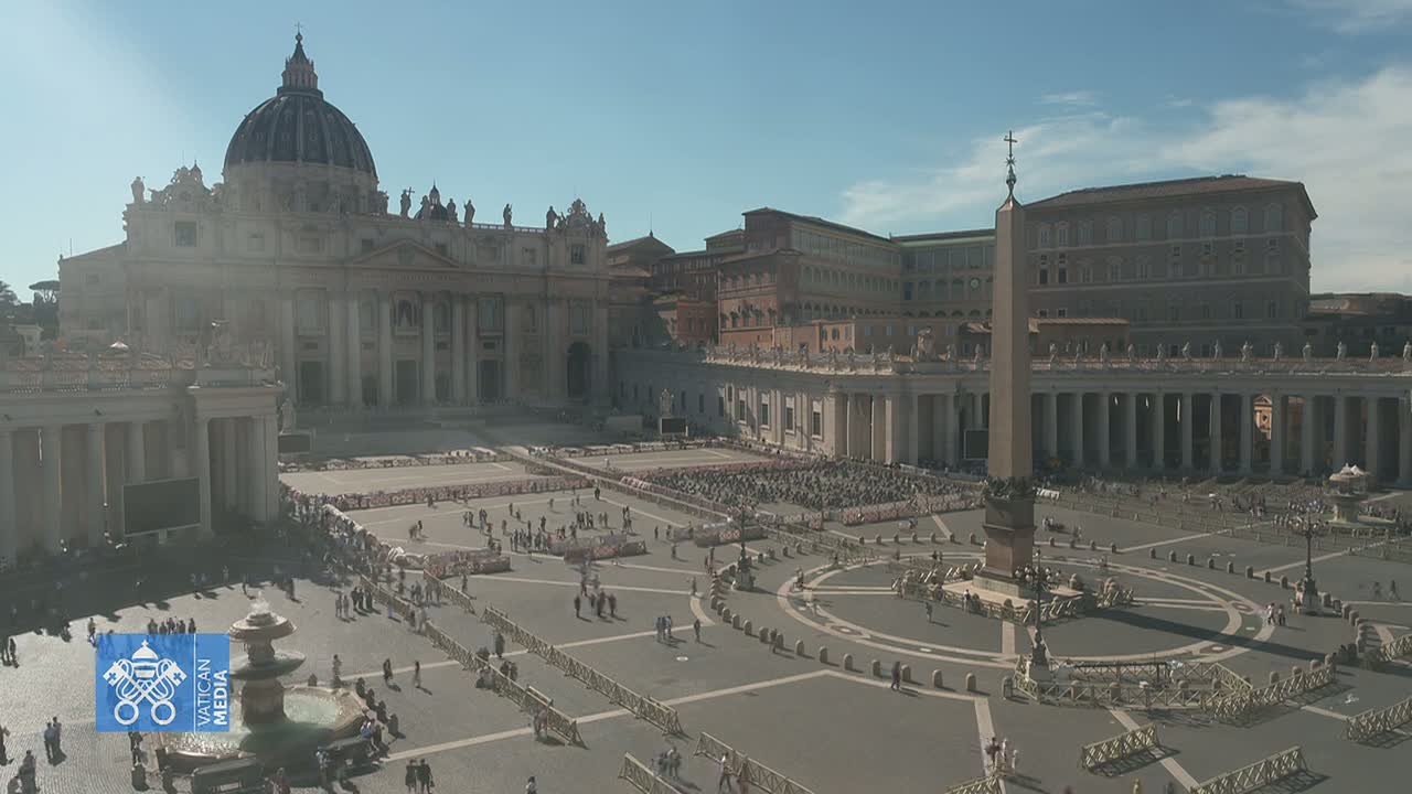 A large crowd gathers in St. Peter's Square, under a bright, sunny sky, in front of St. Peter's Basilica and the obelisk.