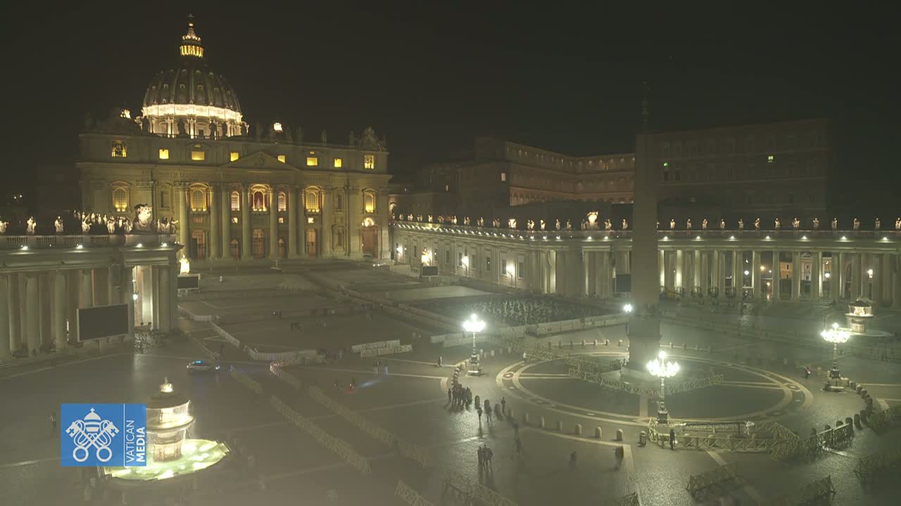 A nighttime, aerial view of St. Peter's Basilica and Square in Vatican City, showing sparsely populated areas with lampposts illuminating the plaza.
