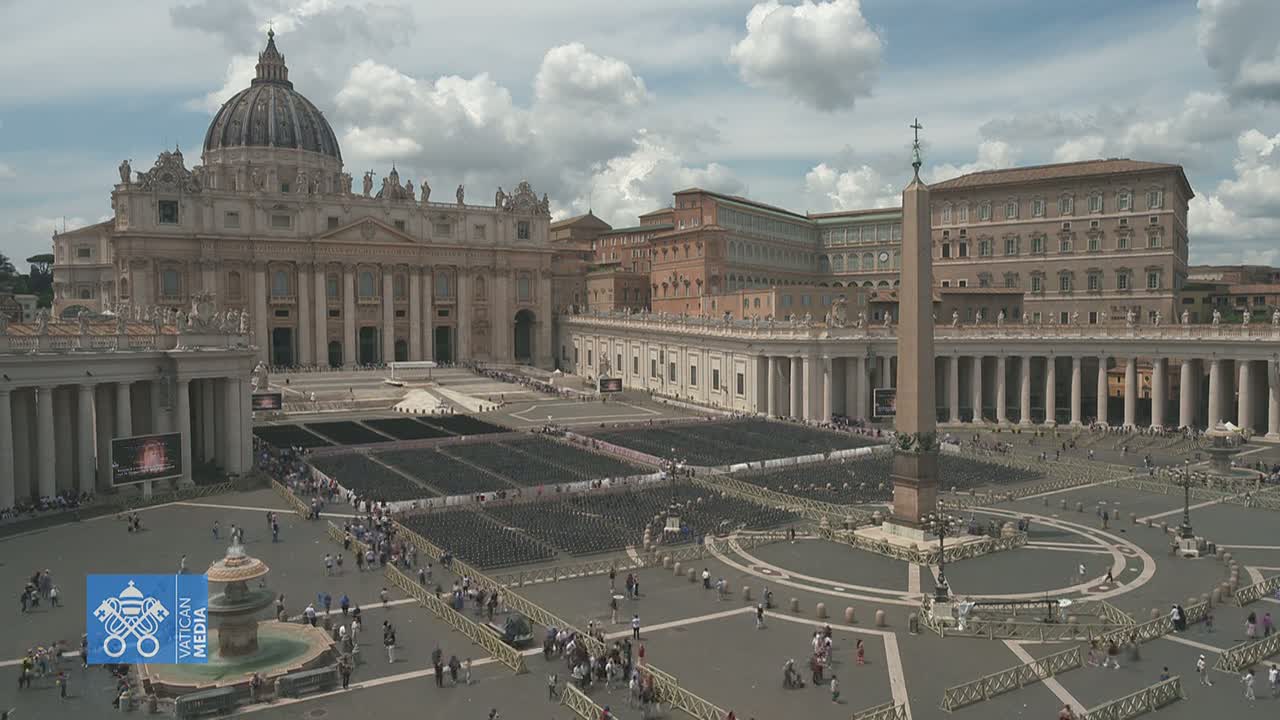 St. Peter's Basilica and St. Peter's Square in Vatican City are shown on a partly cloudy day with many people gathered in the square, which is filled with rows of chairs.