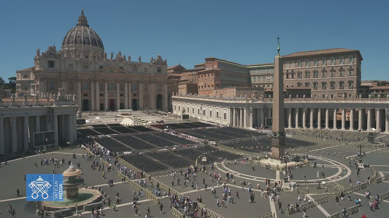 St. Peter's Basilica and St. Peter's Square in Vatican City are shown on a sunny day with many people gathered in the square, rows of chairs set up in the plaza.