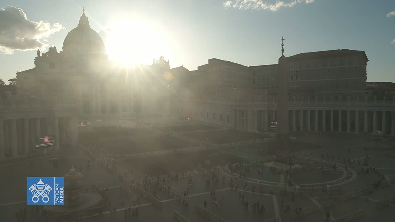 A hazy sun shines brightly behind St. Peter's Basilica in a large, populated square.