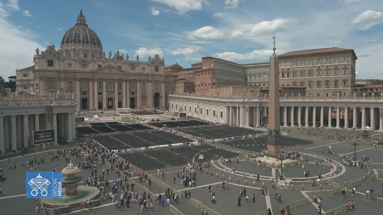 A large crowd gathers in St. Peter's Square in Vatican City, under a partly cloudy sky, with many empty chairs set up in the square in front of St. Peter's Basilica.