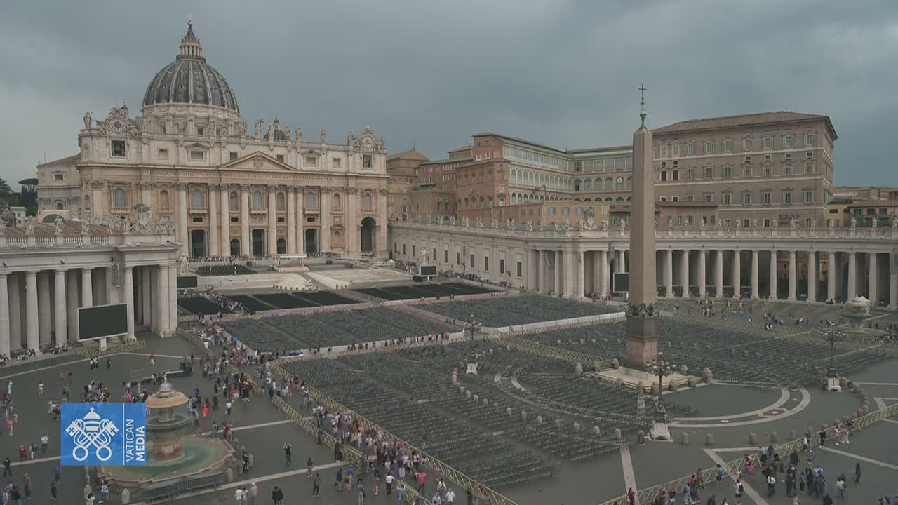 St. Peter's Basilica and St. Peter's Square, filled with many people and rows of empty chairs, under a cloudy sky.