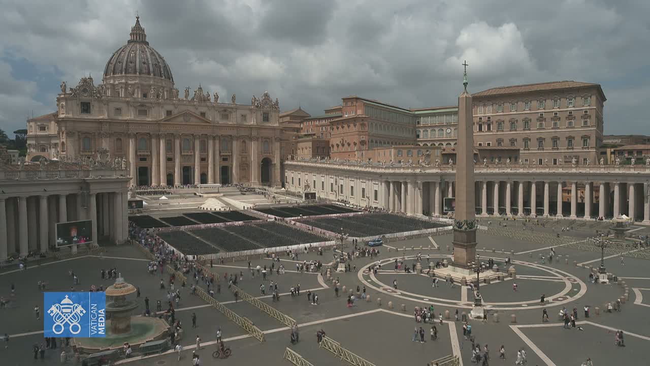 St. Peter's Square in Vatican City, under a cloudy sky, shows a large crowd of people gathered in front of St. Peter's Basilica.