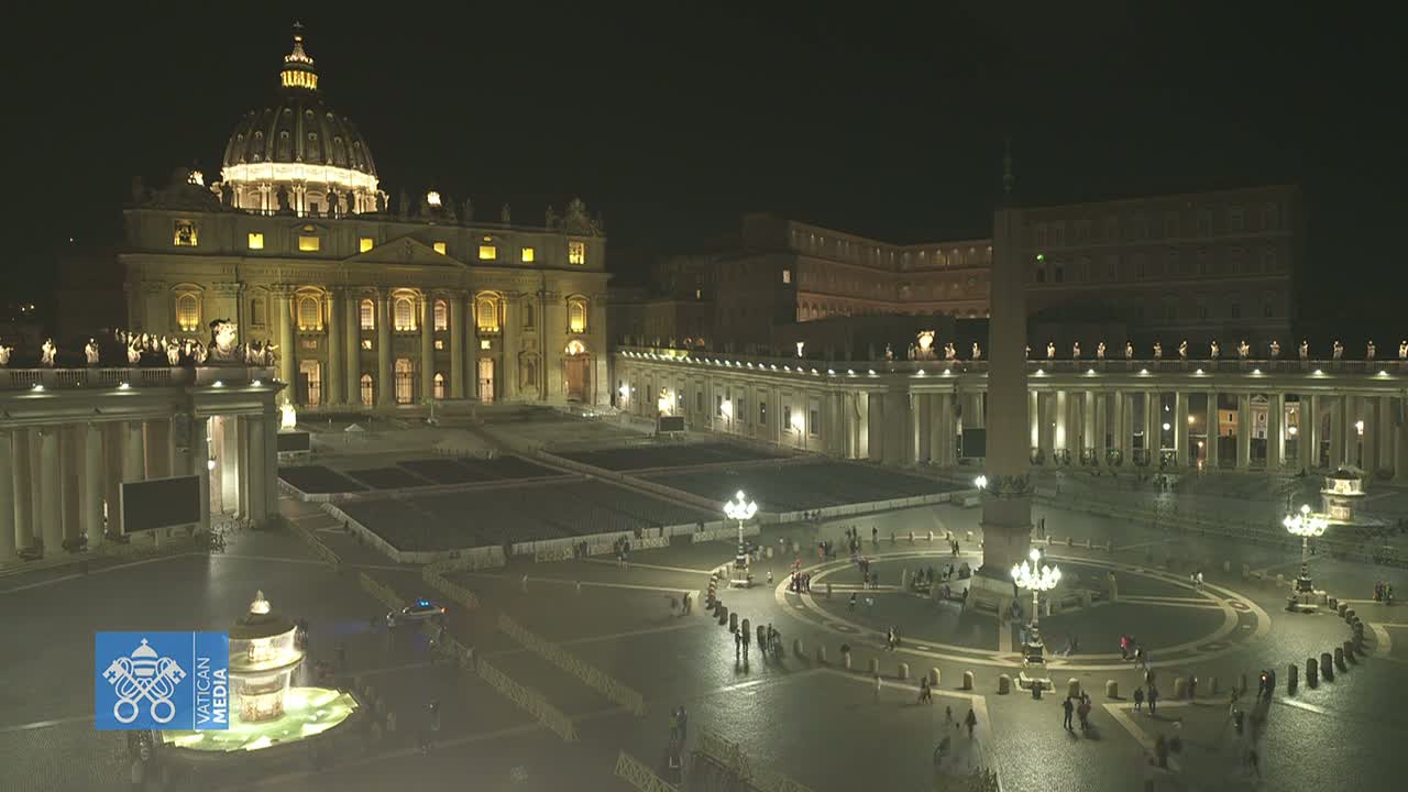 A nighttime, illuminated view of St. Peter's Basilica and St. Peter's Square in Vatican City, with a few people scattered throughout the square.