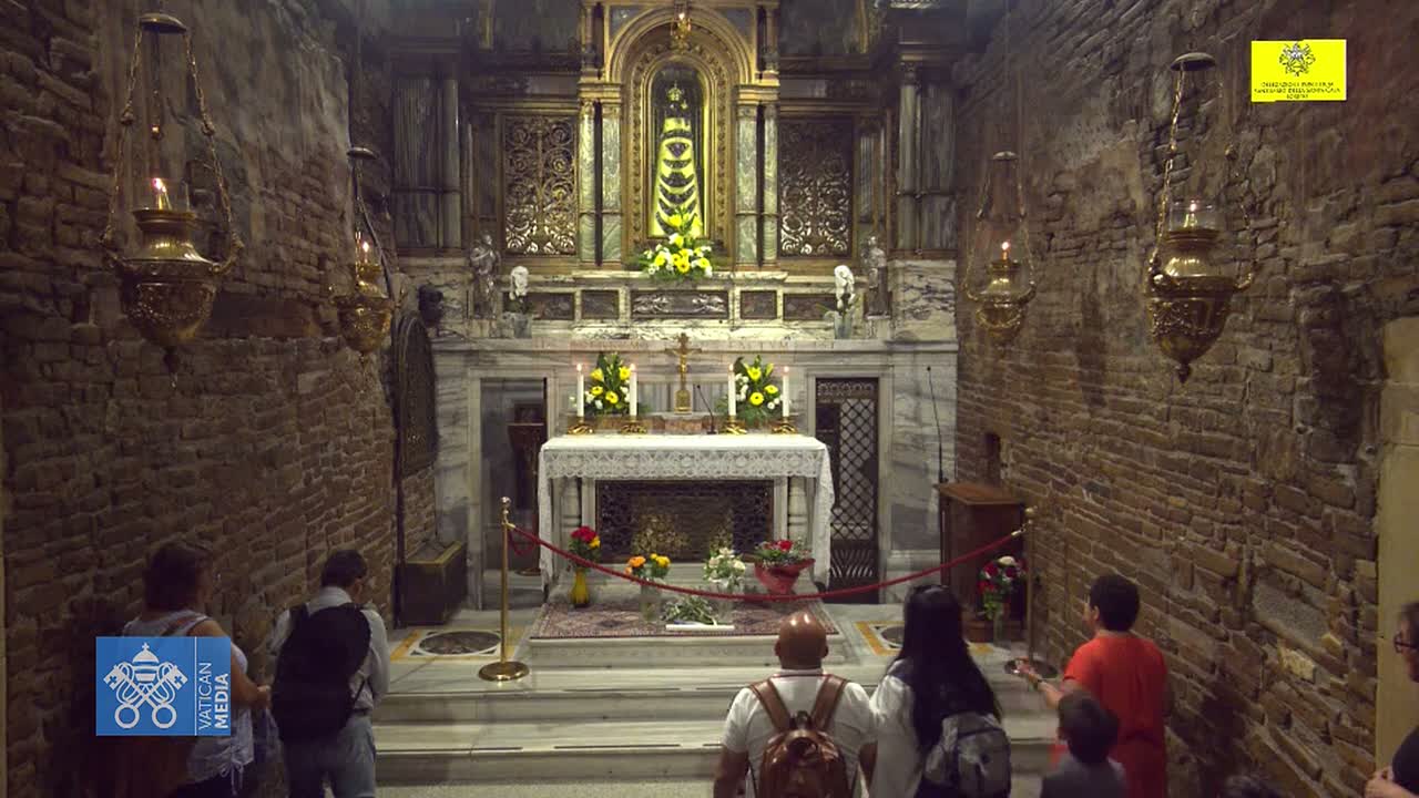 A group of people stand in a dimly lit, brick-walled chapel, observing an altar with religious artifacts and floral arrangements.