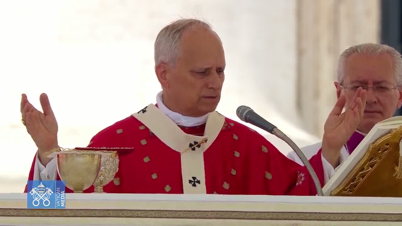 A cardinal in a red robe gestures with his hands while speaking into a microphone, with another man in a clerical robe assisting him.