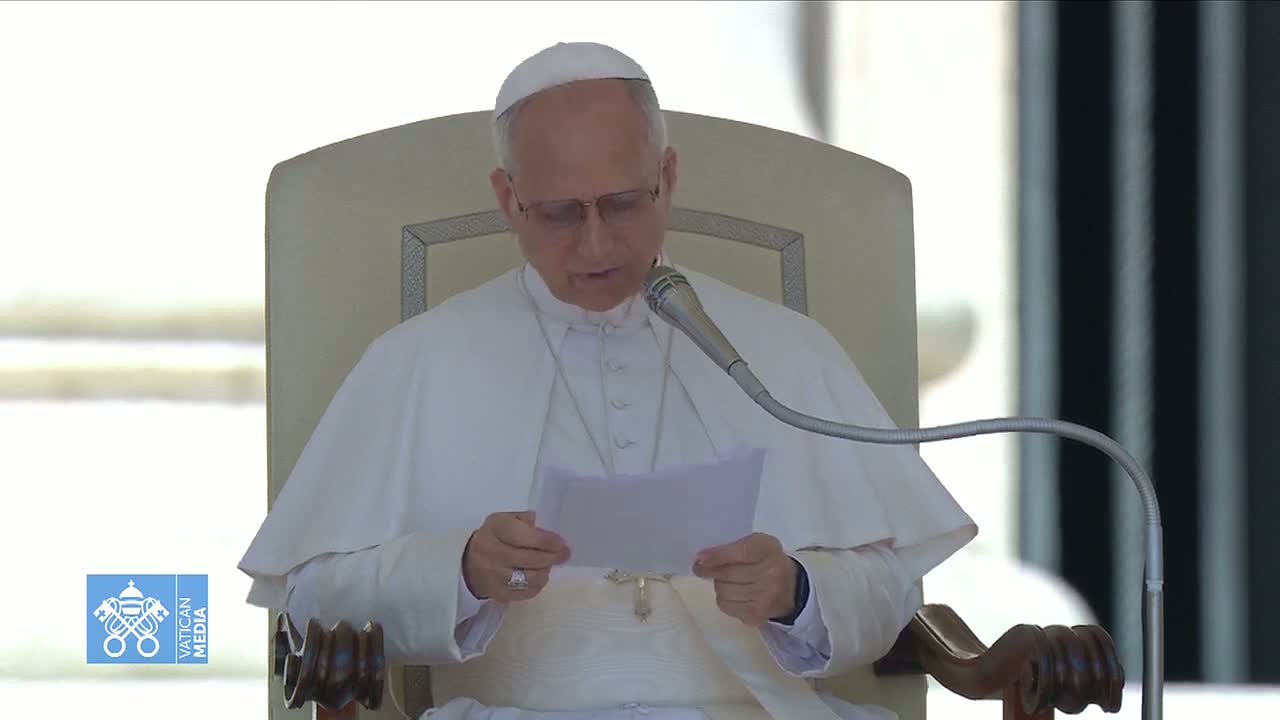 The Pope, wearing a white cassock, sits in a chair and reads from a piece of paper at a microphone.