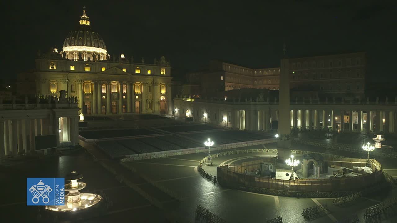 St. Peter's Basilica and a mostly empty St. Peter's Square are brightly illuminated at night, with the damp ground reflecting lights under a dark sky.