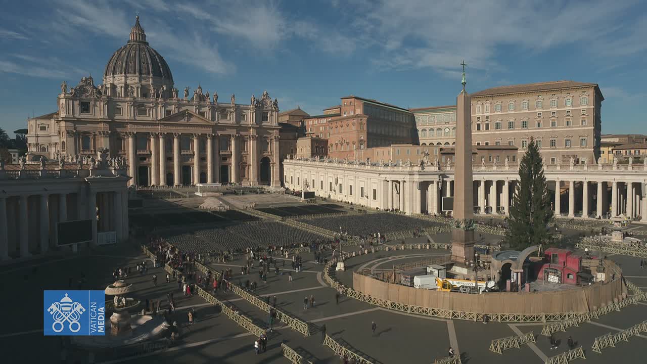 St. Peter's Square is seen on a sunny day with a blue sky and scattered clouds, featuring St. Peter's Basilica, an obelisk next to a large Christmas tree being prepared for display, and extensive rows of empty seating set up for an event, with many people walking across the square.