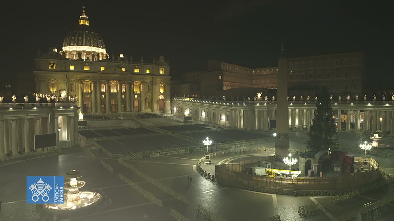 St. Peter's Square is seen on a sunny day with a blue sky and scattered clouds, featuring St. Peter's Basilica, an obelisk next to a large Christmas tree being prepared for display, and extensive rows of empty seating set up for an event, with many people walking across the square.