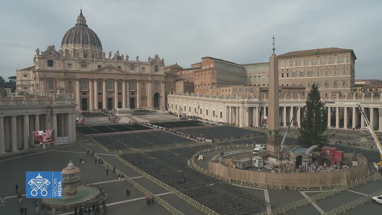 St. Peter's Square is seen on an overcast day, featuring St. Peter's Basilica, the Vatican Obelisk, a large Christmas tree, and a nativity scene under construction, all surrounded by rows of empty chairs.