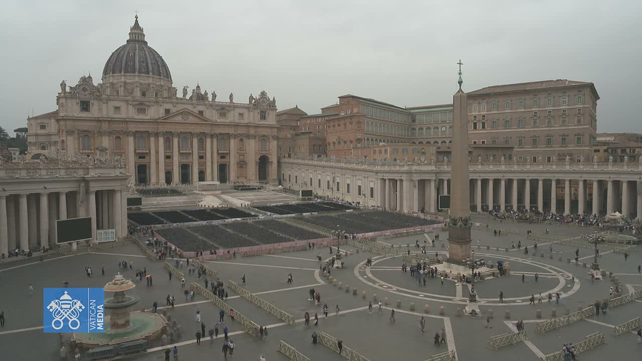 St. Peter's Square is seen on an overcast day, featuring St. Peter's Basilica, the Vatican Obelisk, a large Christmas tree, and a nativity scene under construction, all surrounded by rows of empty chairs.