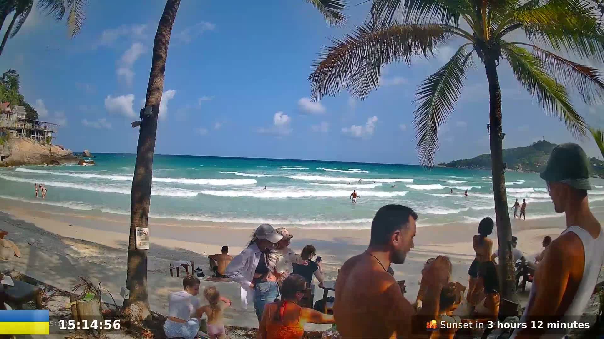 A sunny beach scene shows people relaxing and swimming in turquoise water with gentle waves under a mostly clear sky.