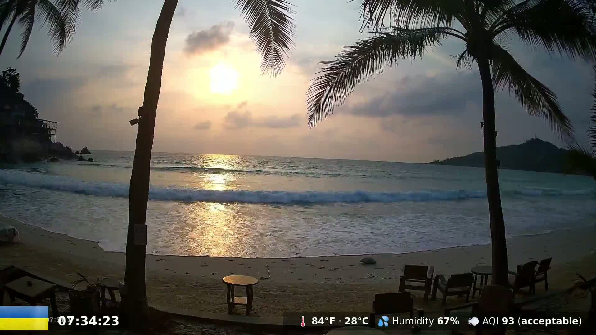 A beach at sunset, with gentle waves lapping the shore, palm trees framing the view, and outdoor furniture on the sand.