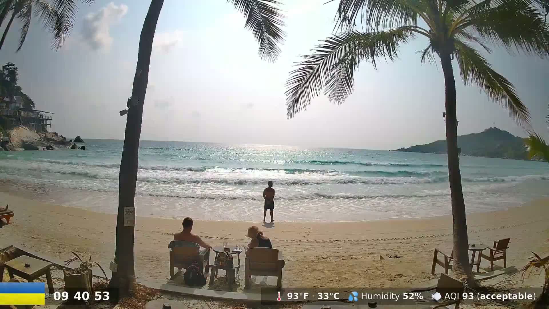 A sandy beach with gentle waves laps the shore under a sunny sky, palm trees framing the scene as people relax in beach chairs.