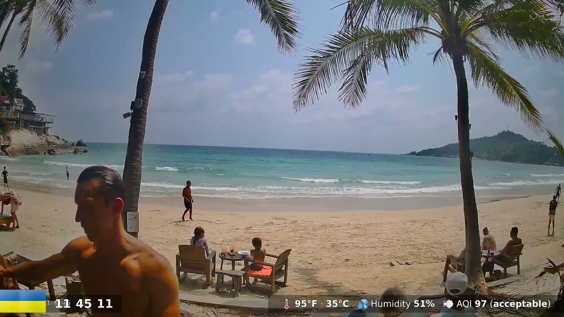 A sandy beach with turquoise water, palm trees, and several people relaxing on the shore under a partly sunny sky.