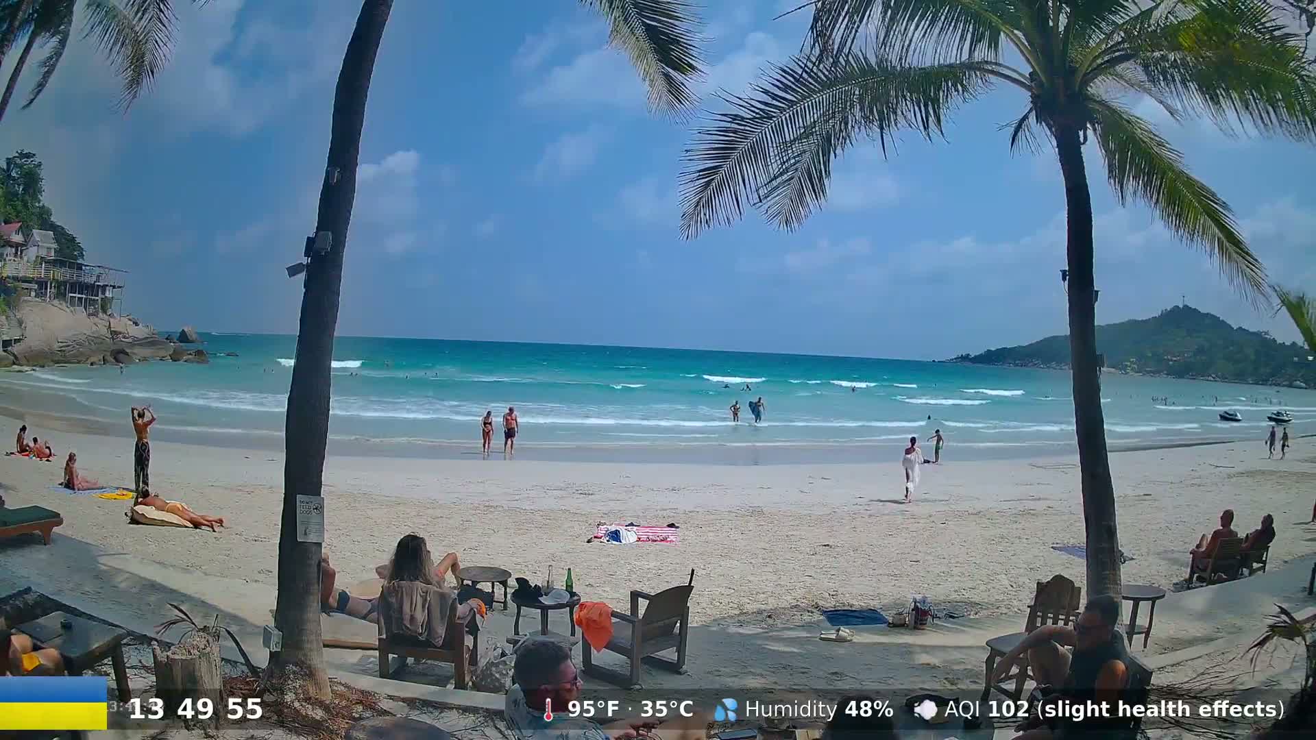 A tropical beach with turquoise water and white sand, under a partly cloudy sky, features several people swimming, sunbathing, and relaxing on beach chairs near palm trees.