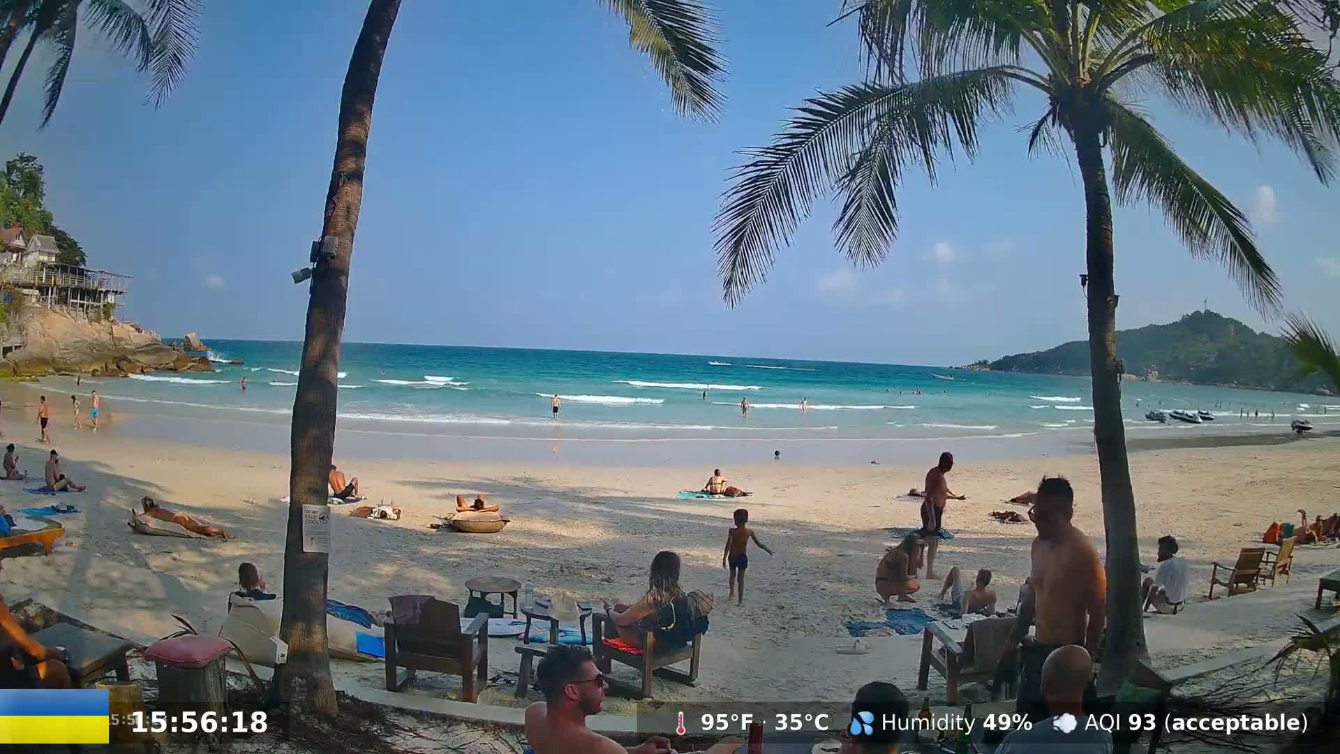 A sunny beach scene shows people relaxing and swimming in the ocean under a clear blue sky.
