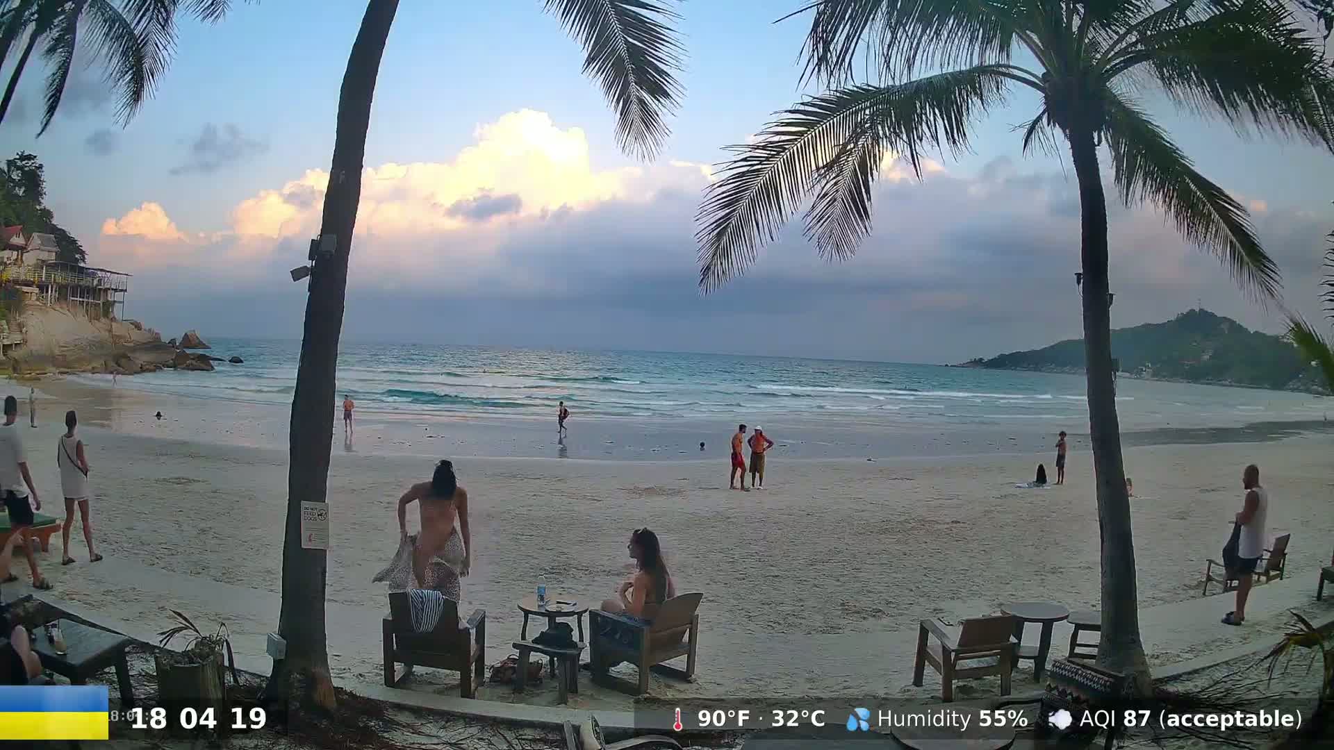 A sandy beach with palm trees and several people, some swimming in the ocean under a partly cloudy sky.