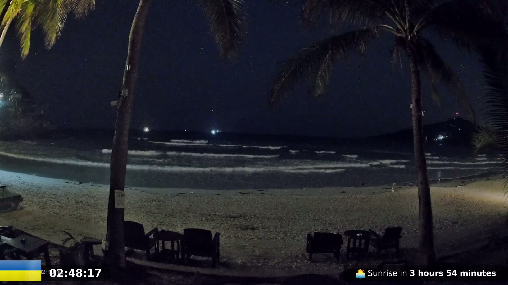 A nighttime beach scene under a starry sky shows gentle waves lapping the shore, with palm trees and outdoor furniture in the foreground.